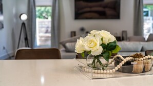 Una habitación con una mesa de vidrio transparente que exhibe rosas blancas, un collar de cuentas y un cuenco de madera.