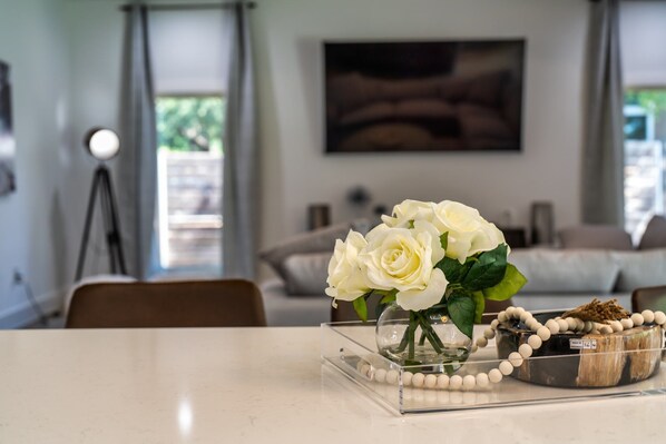 A room with a clear glass table displaying white roses, a beaded necklace, and a wooden bowl. - The Gateway 6BED 2 Houses GameRoom Pool (Cedar Park)