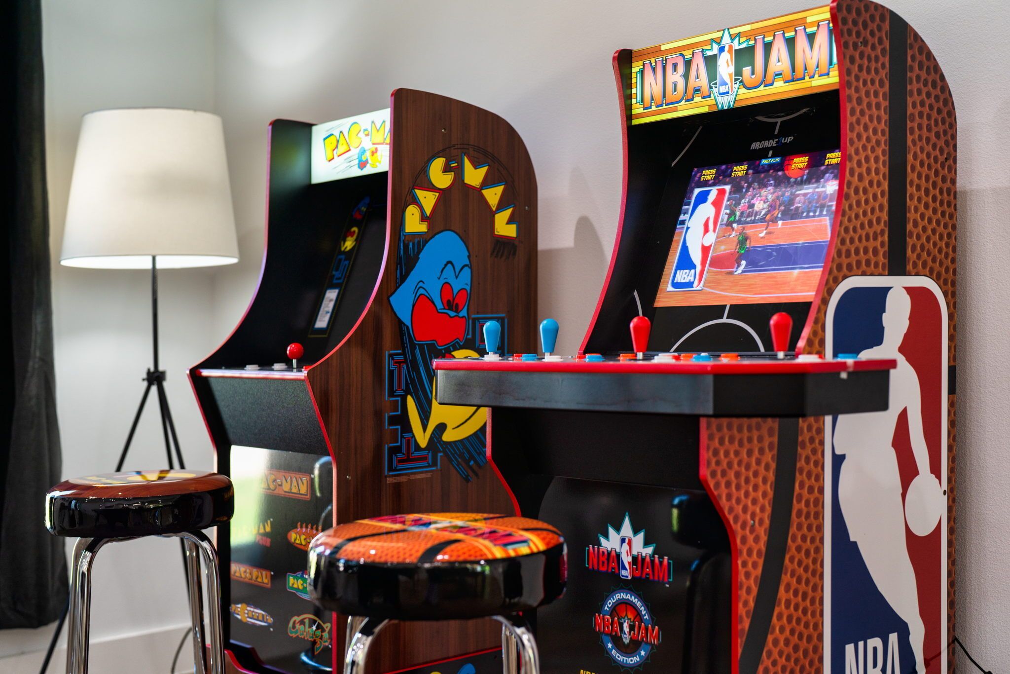 Arcade machines with Pac-Man and NBA Jam games, a lamp, and a stool.