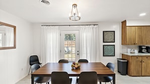 Un comedor con una mesa de madera, sillas negras y un frutero. Se observa una cocina con armarios de madera y un lavamanos.