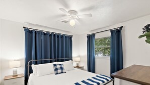 Un dormitorio con cama, dos mesitas de noche, un velador, ventilador de techo y ventana con cortinas.