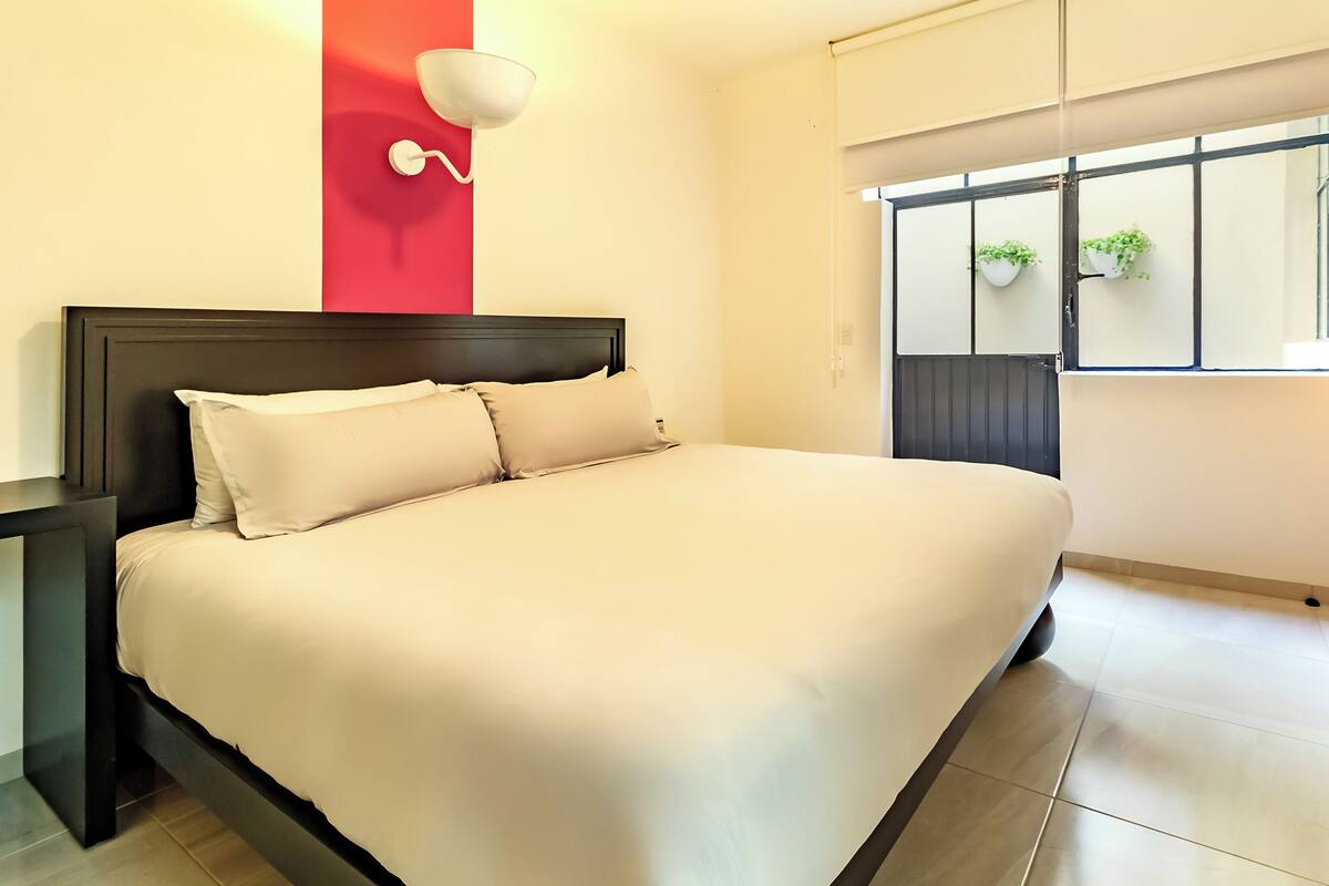 A modern bedroom with a large bed, a nightstand, a red accent wall, and a window with plants.