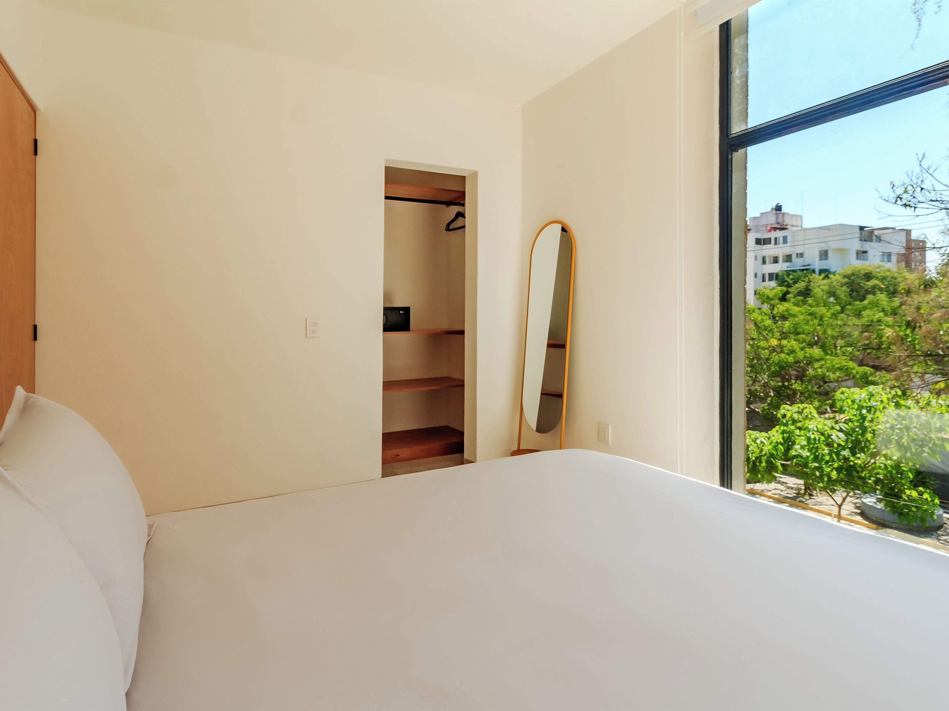 A bedroom with a bed, a wardrobe, a mirror, and a window with a view of trees and buildings.