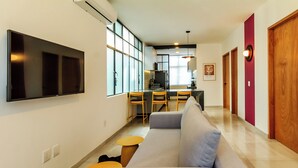 Ein modernes Wohnzimmer mit grauem Sofa, einem Flachbildfernseher und einem Essbereich mit Holzsesseln und einem kleinen Tisch.