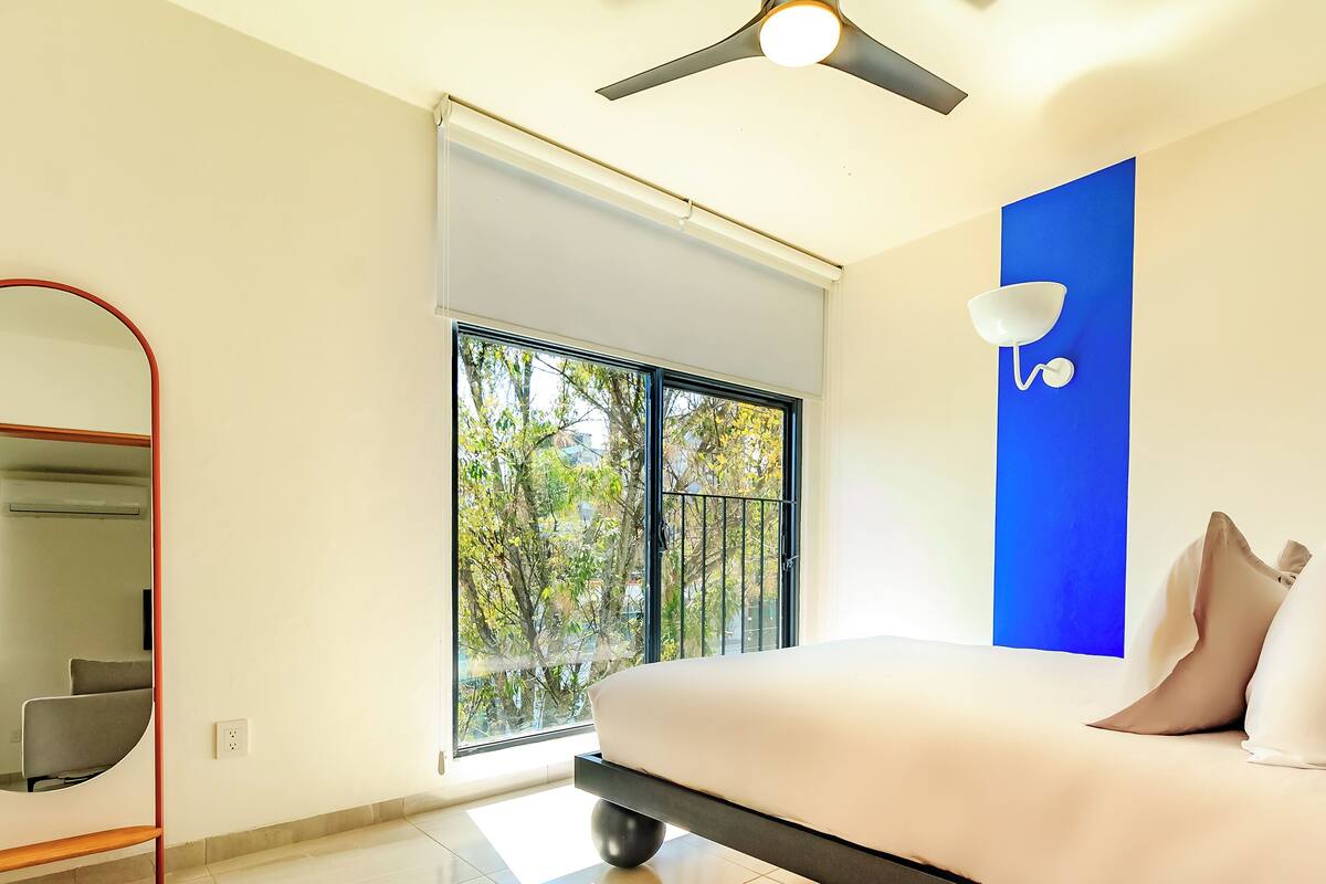 A modern bedroom with a large window, a ceiling fan, and a bed with pillows.