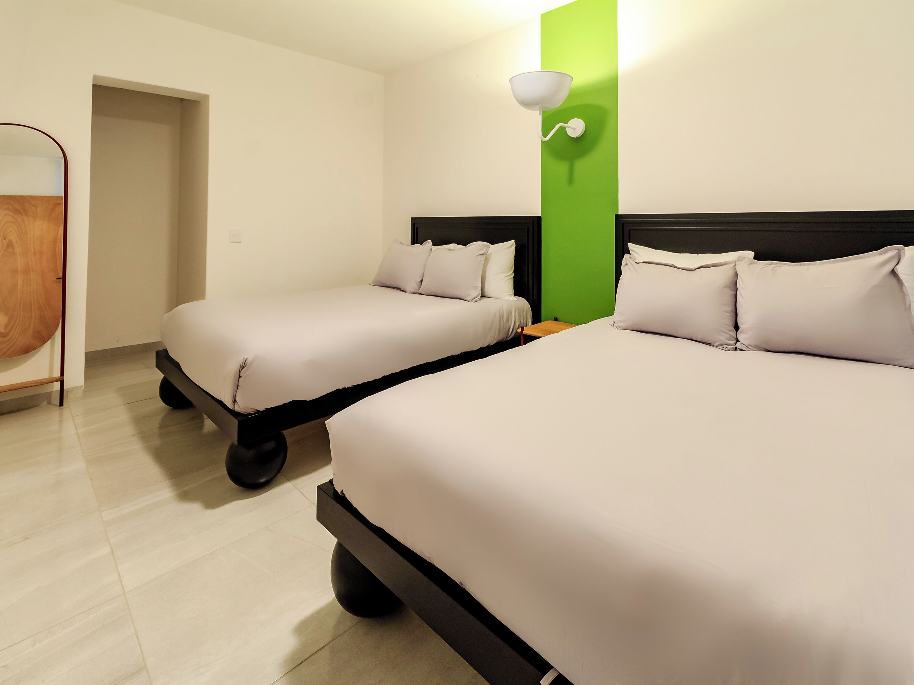 A hotel room with two beds, a green accent wall, and a bedside table with a lamp.