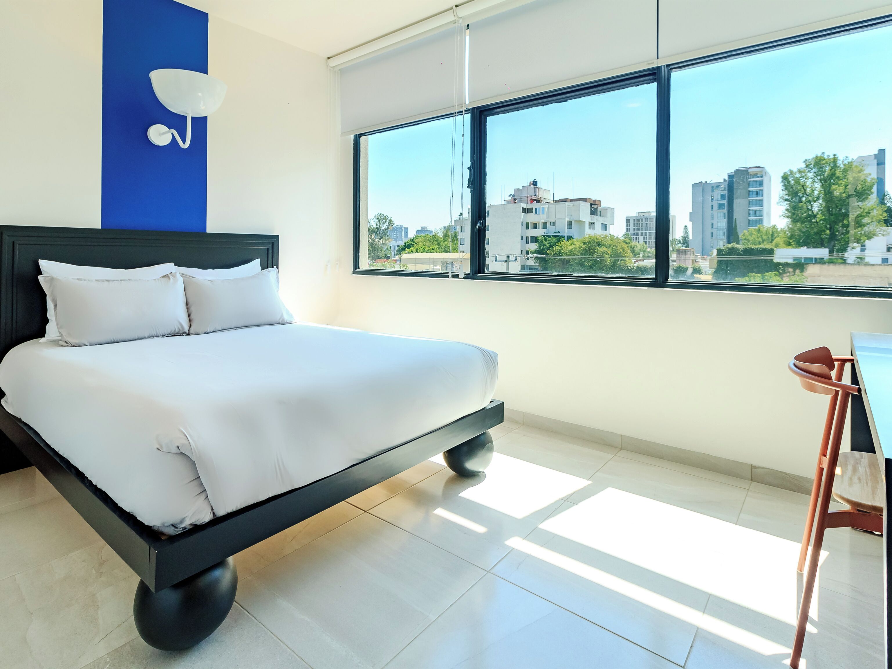 Una habitación de hotel moderna con una cama grande, vistas de la ciudad a través de un ventanal amplio y un escritorio con una silla.