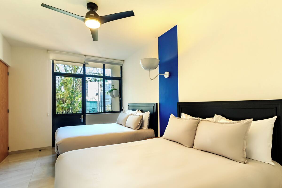 A hotel room with two beds, a ceiling fan, a window with a view of trees, and a wall-mounted light fixture.