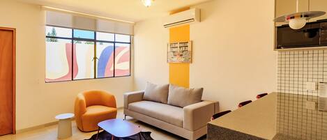 A modern living room with a sofa, armchair, and a coffee table. There is a large window with abstract art, an air conditioning unit, and a kitchen area with a tiled backsplash.