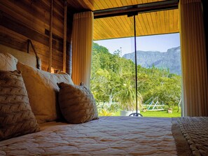 A bed with a quilted cover and two pillows, a wooden ceiling, and a view of a lush green landscape with mountains in the distance. - Vila Vicença Natural Refuge (Praia Grande)