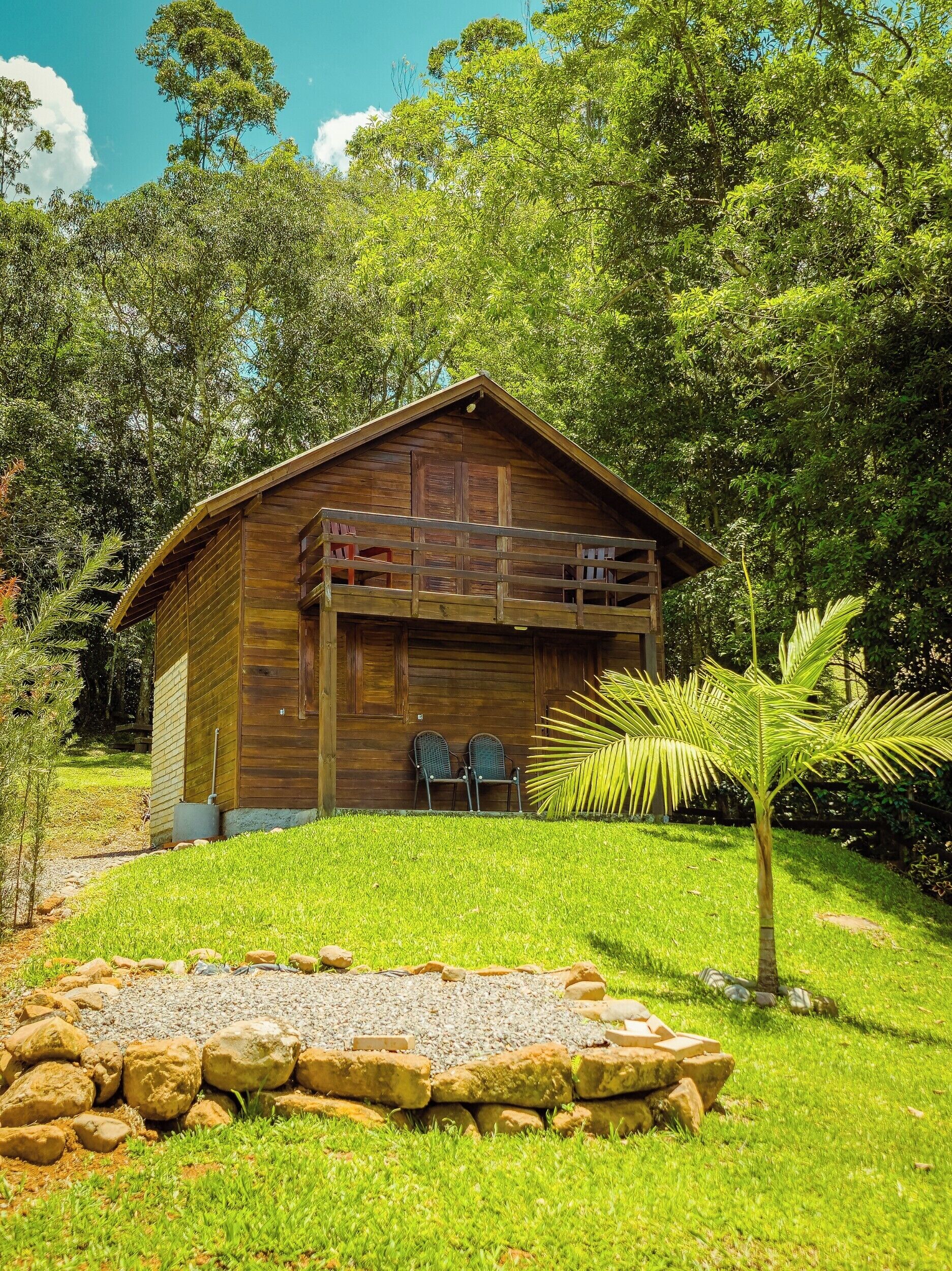 A wooden cabin with a balcony, surrounded by lush greenery and a small palm tree.