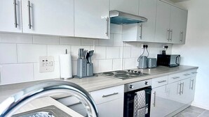 Μια σύγχρονη κουζίνα με λευκά ντουλάπια, έναν απορροφητήρα από ανοξείδωτο ατσάλι, έναν εντοιχισμένο φούρνο και έναν πάγκο εργασίας με βραστήρα και φρυγανιέρα.