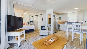 Un salon moderne et bien éclairé, doté d’un téléviseur à écran plat, d’une table basse en bois et d’un coin repas avec des chaises blanches.
