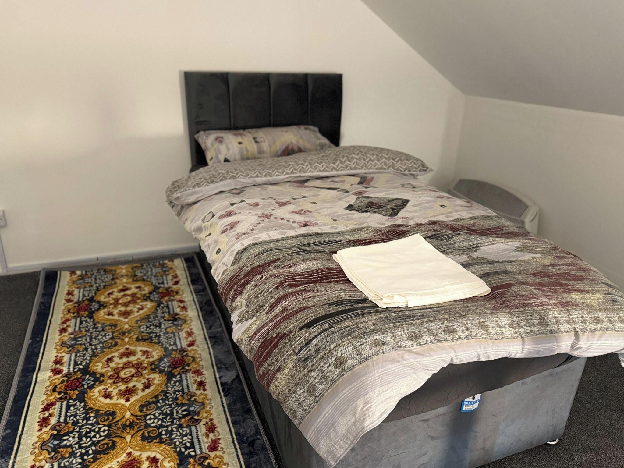 Un lit simple avec une couvre-lit à motifs et des oreillers, une tête de lit grise et un tapis décoratif sur le sol.