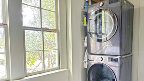 A laundry room with a stacked washer and dryer, a window with a view of trees, and a bed with a white quilt. - Charming Home 2 miles to UGA (Athens)