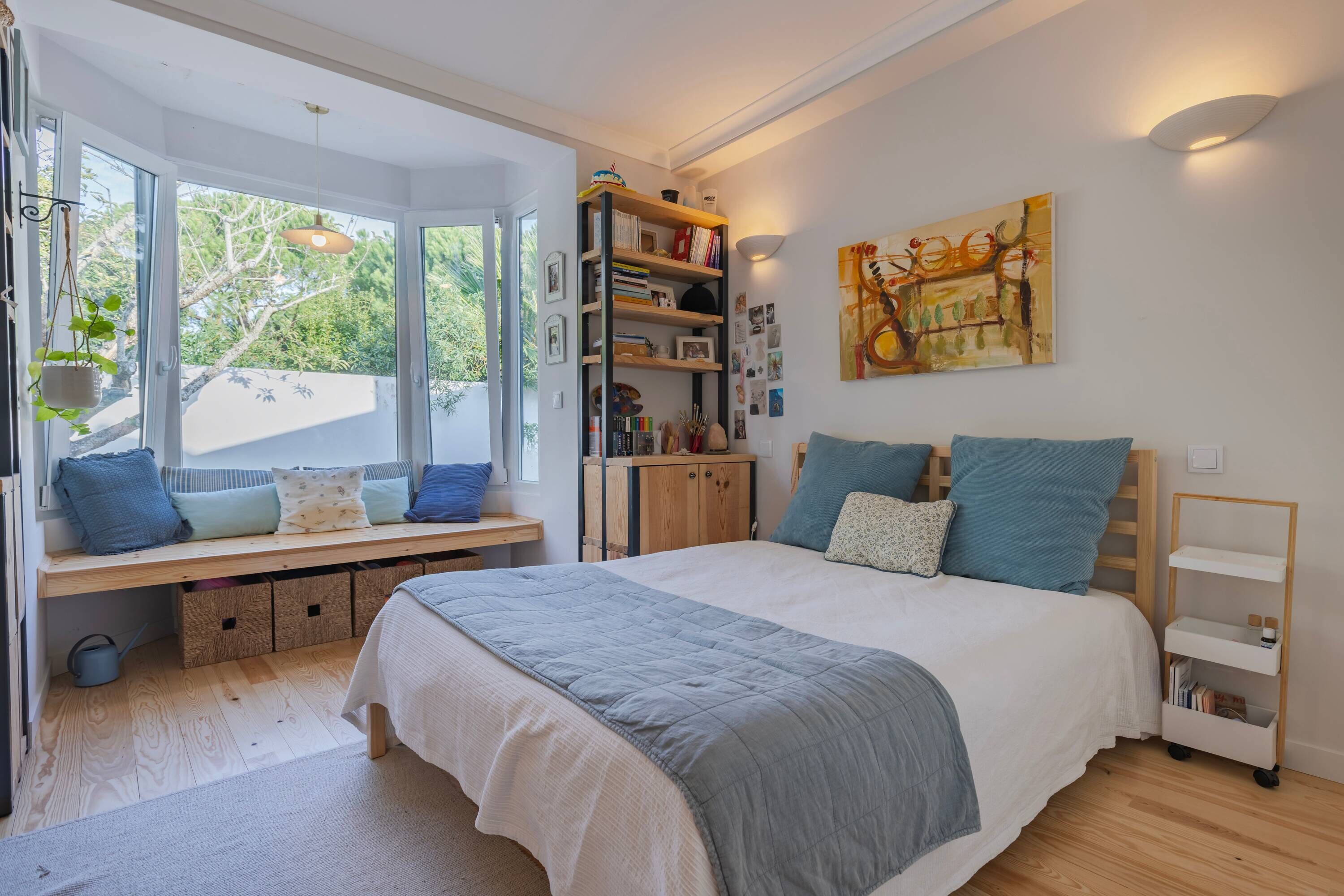 Un dormitorio con una cama grande, un banco empotrado con espacio de almacenamiento, una estantería y una ventana con vistas a árboles.