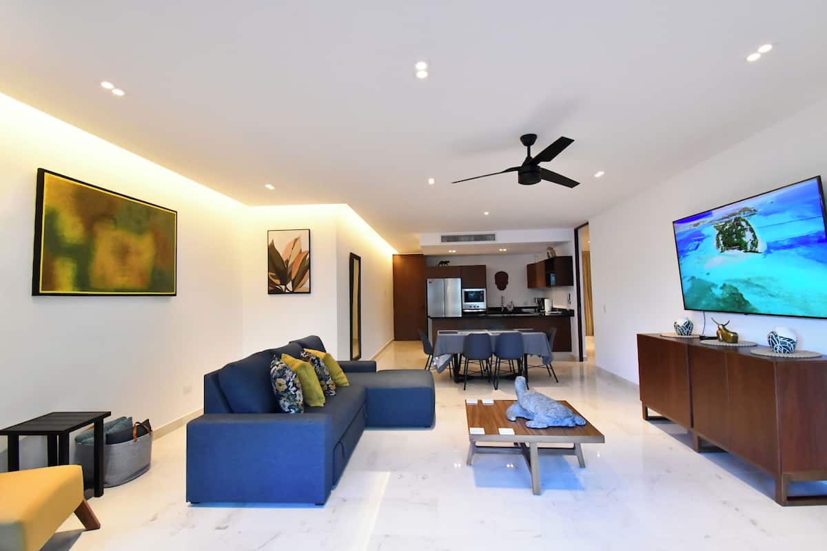A modern living room with a blue sofa, a flat-screen TV, and a dining area in the background.