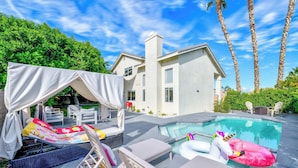 Un espace piscine avec des chaises longues, un coin salon abrité et une maison en arrière-plan.