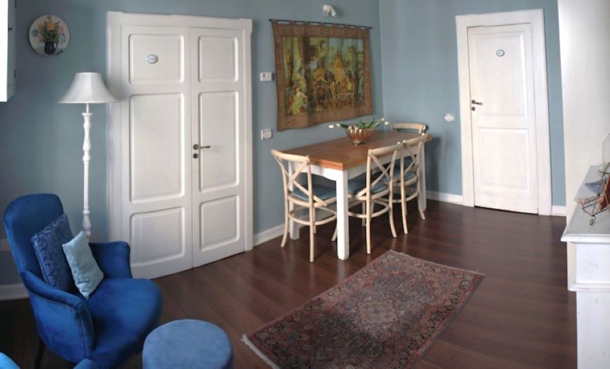 A room with a dining table, chairs, a blue armchair, a white cabinet, and a patterned rug.