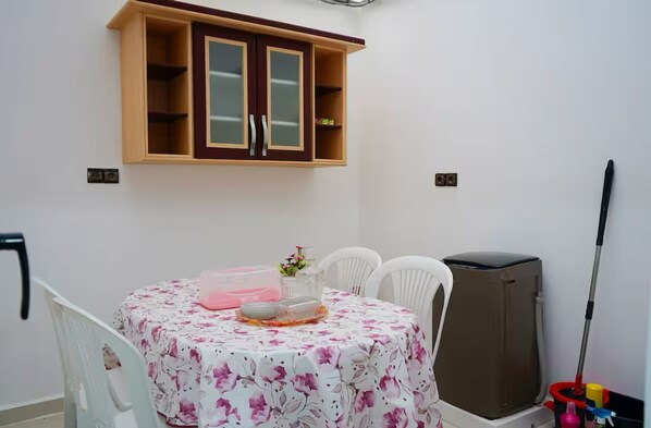A dining area with a table covered in a floral tablecloth, a cabinet above, and a trash bin to the side.