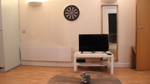 A living room with a flat-screen TV on a white stand, a dartboard on the wall, and a small decorative item on a round table.