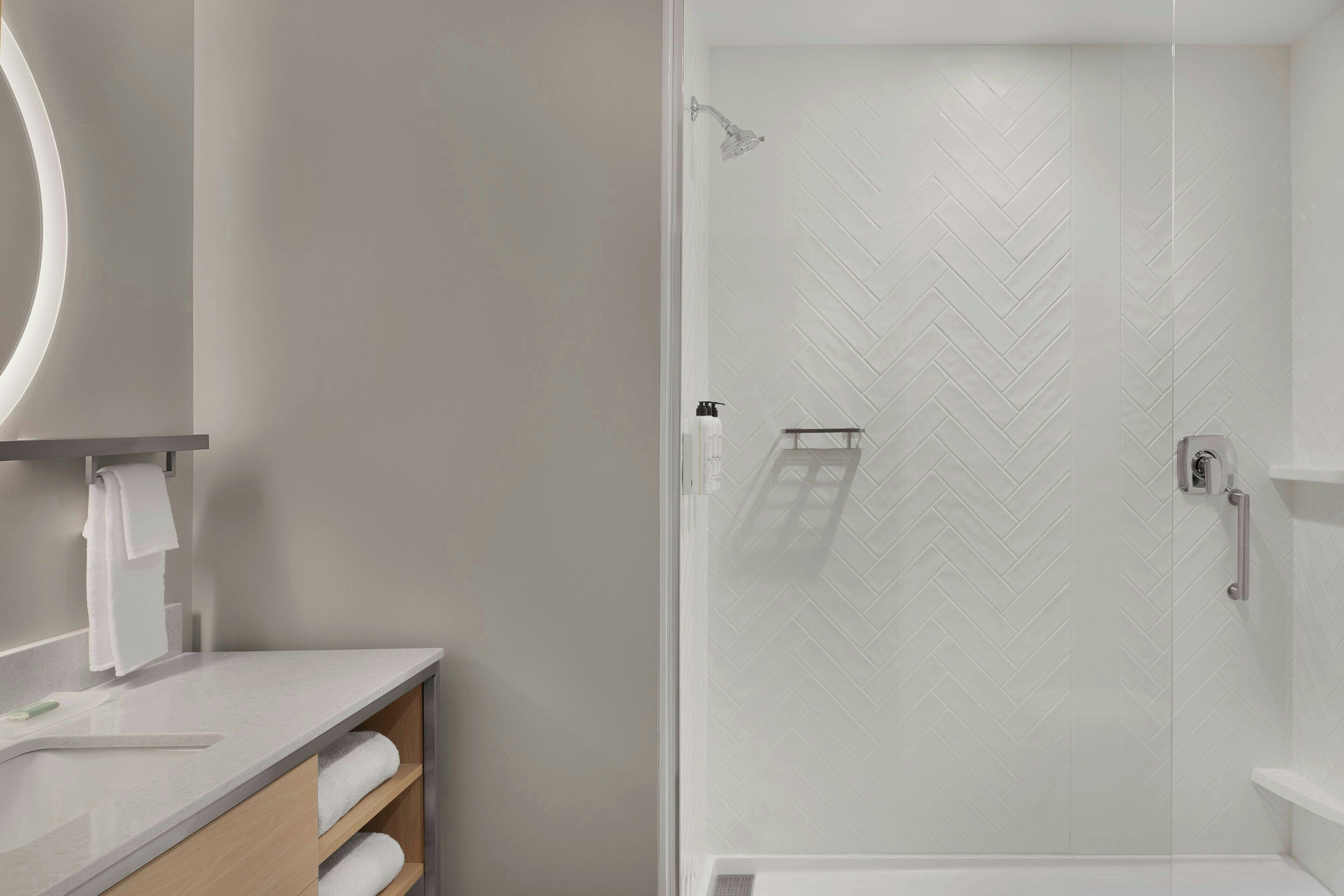 Une salle de bain moderne avec une douche carrelée de motifs en chevrons blancs, un lavabo blanc avec un miroir et une étagère en bois où sont posées des serviettes.