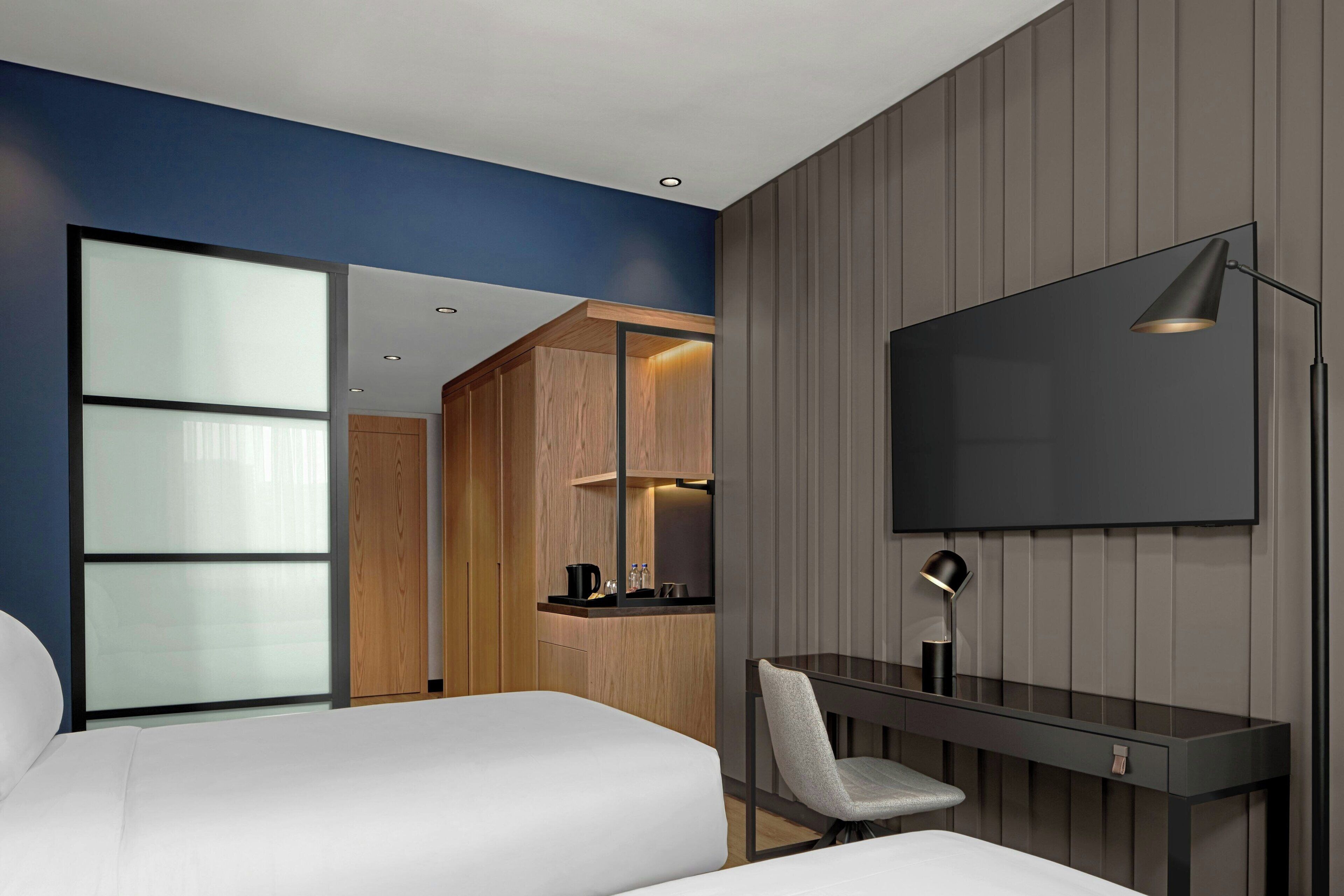 A modern hotel room with a large bed, a desk with a chair, a flat-screen TV mounted on the wall, and a wooden cabinet with a built-in mirror.