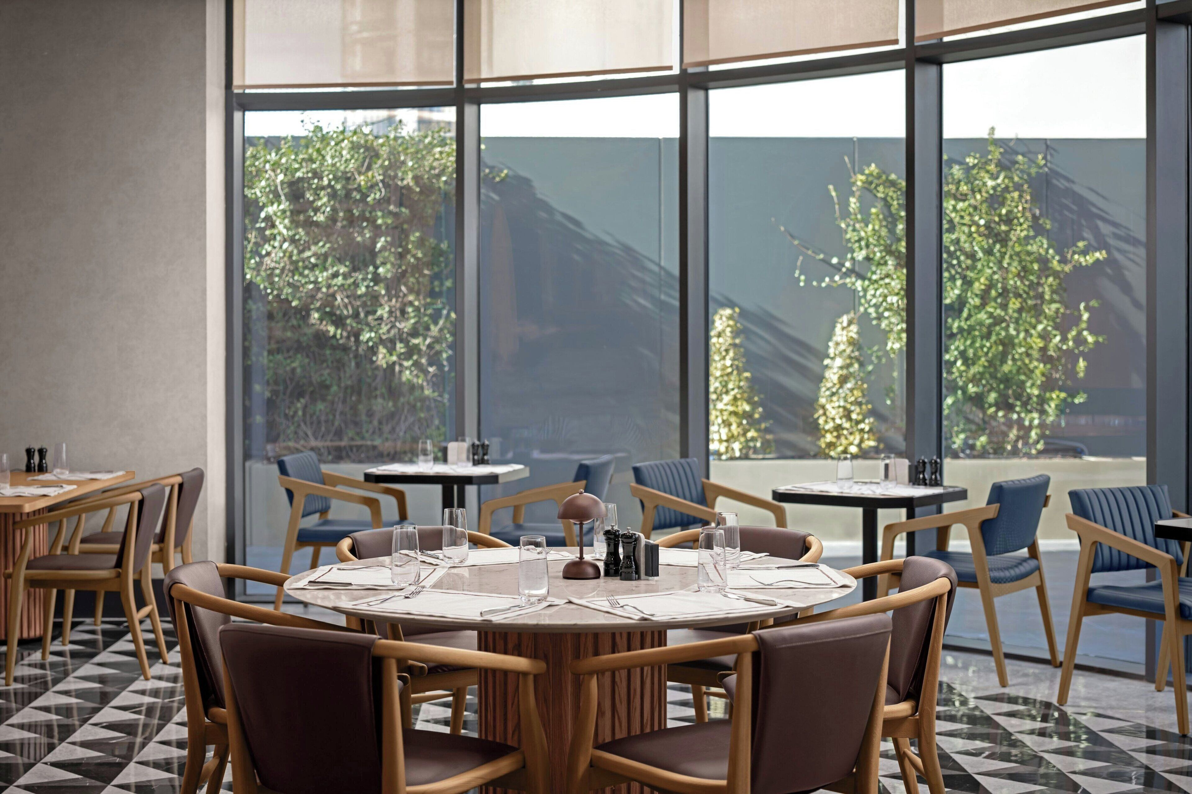 A dining area with round tables, chairs, and a large window offering a view of greenery outside.
