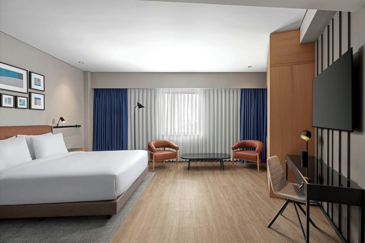 Une chambre d’hôtel avec un grand lit, deux fauteuils, une petite table, un téléviseur et une fenêtre avec des rideaux.