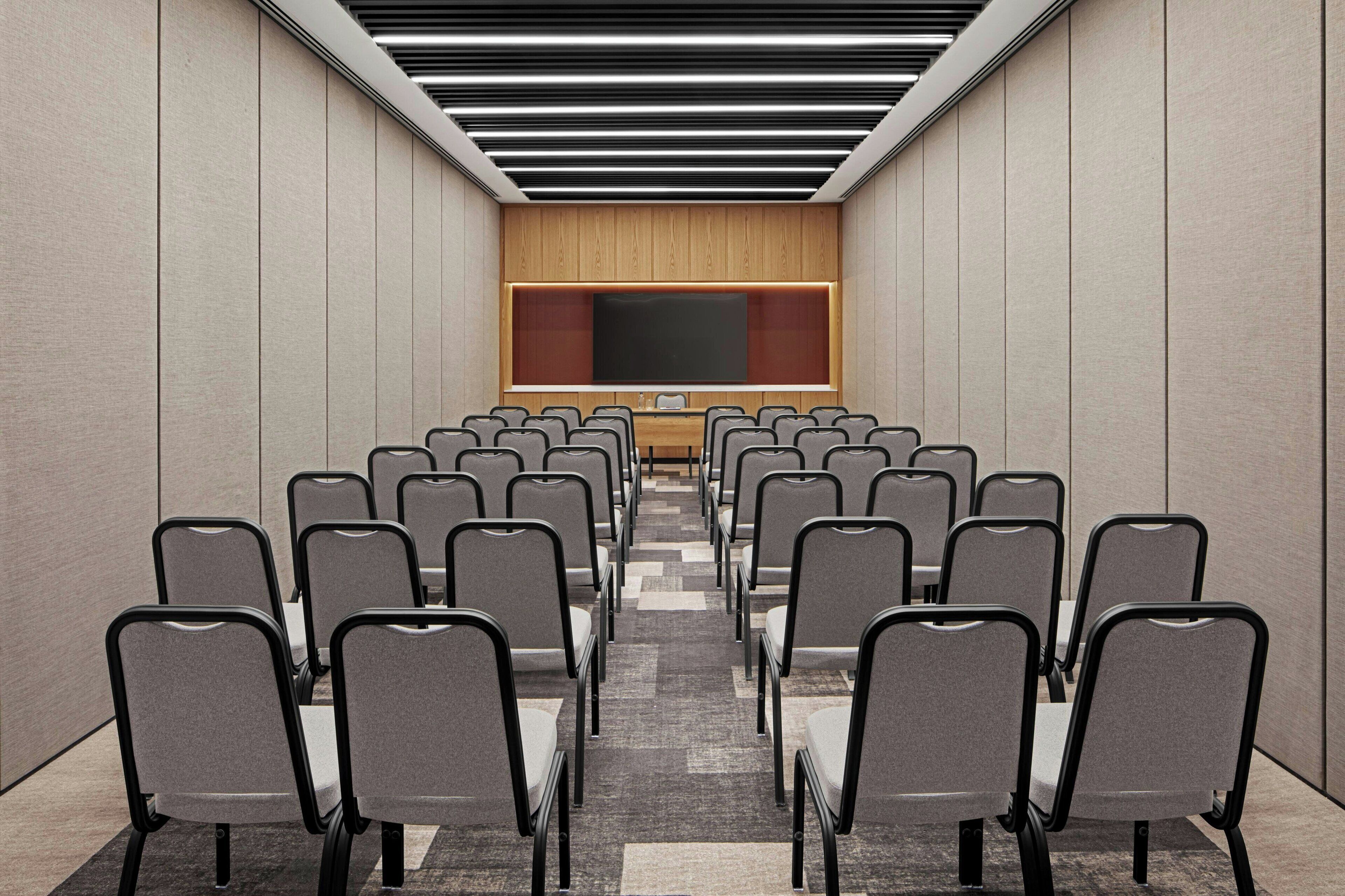 A conference room with rows of chairs facing a large screen, a wooden panel wall, and a patterned carpet.