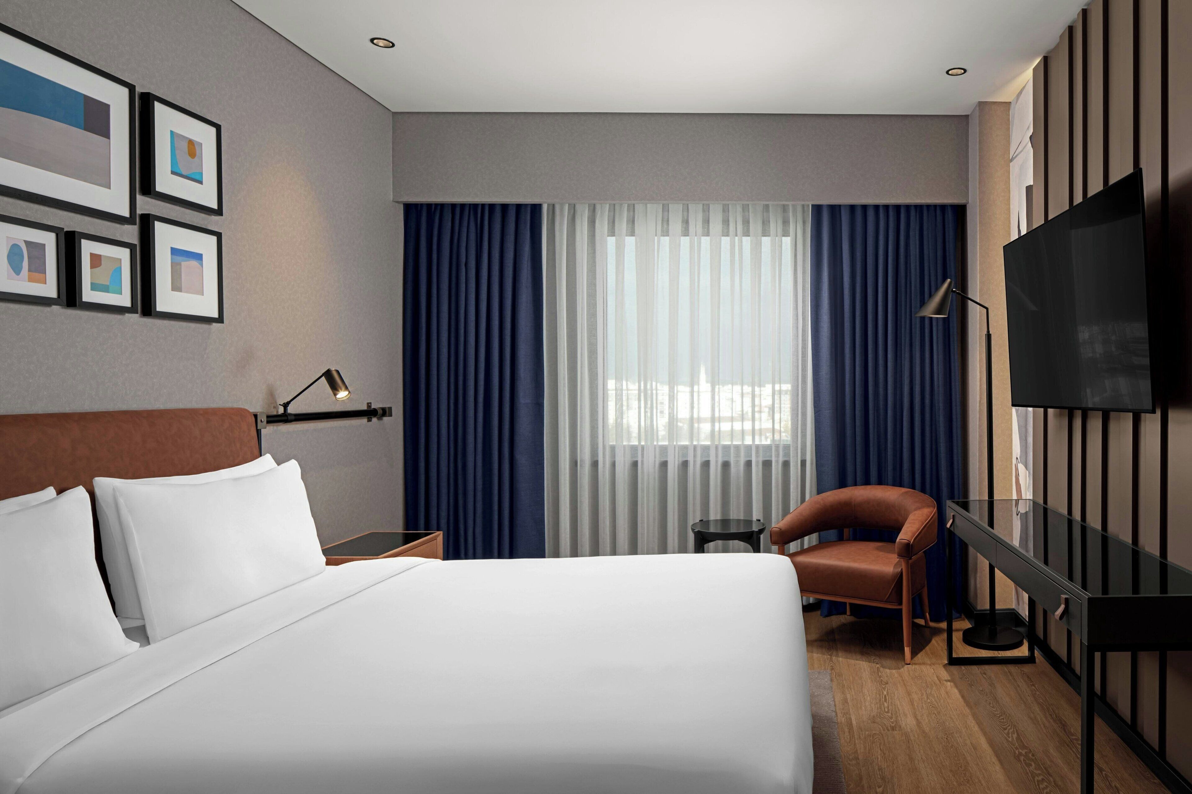 A hotel room with a large bed, a desk with a flat-screen TV, a chair, and a window with curtains.