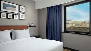 Hotelkamer met een bed, nachtkastje, lamp, stadszicht en lijstjes met kunst aan de muur.