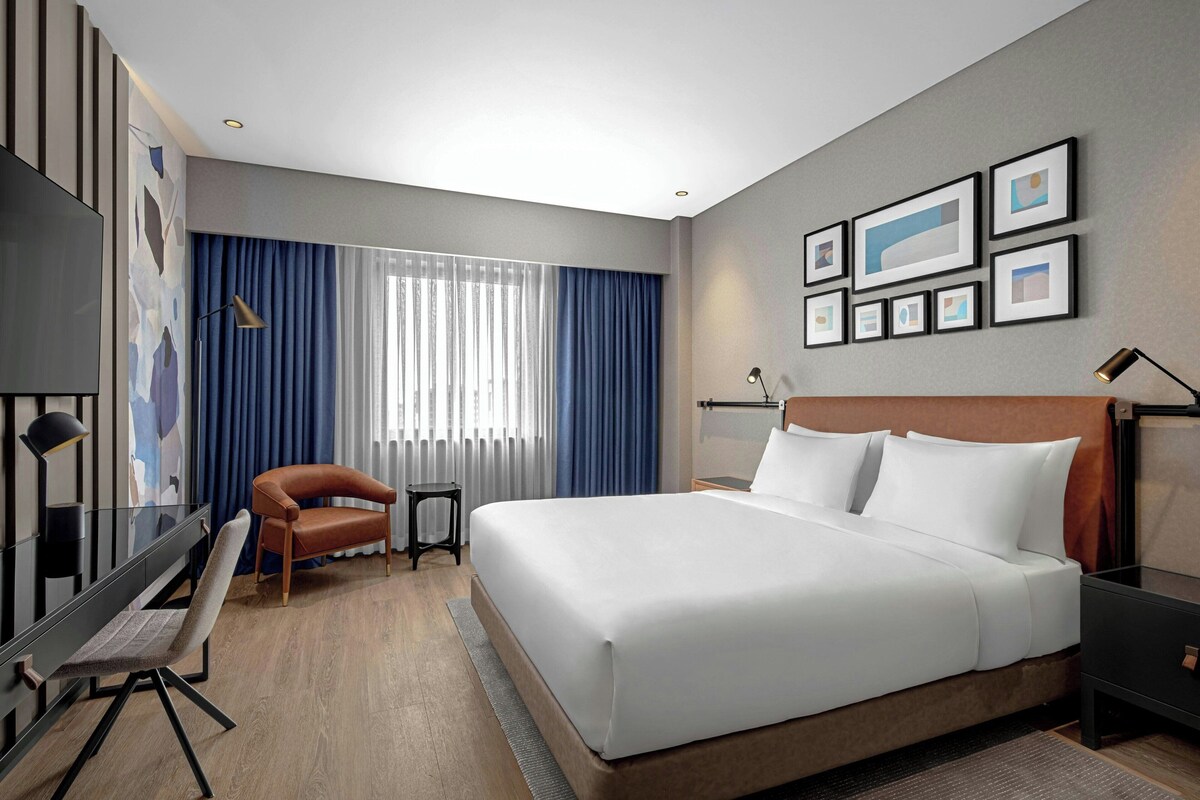 A modern hotel room with a large bed, a desk with a chair, a small table, and a TV.