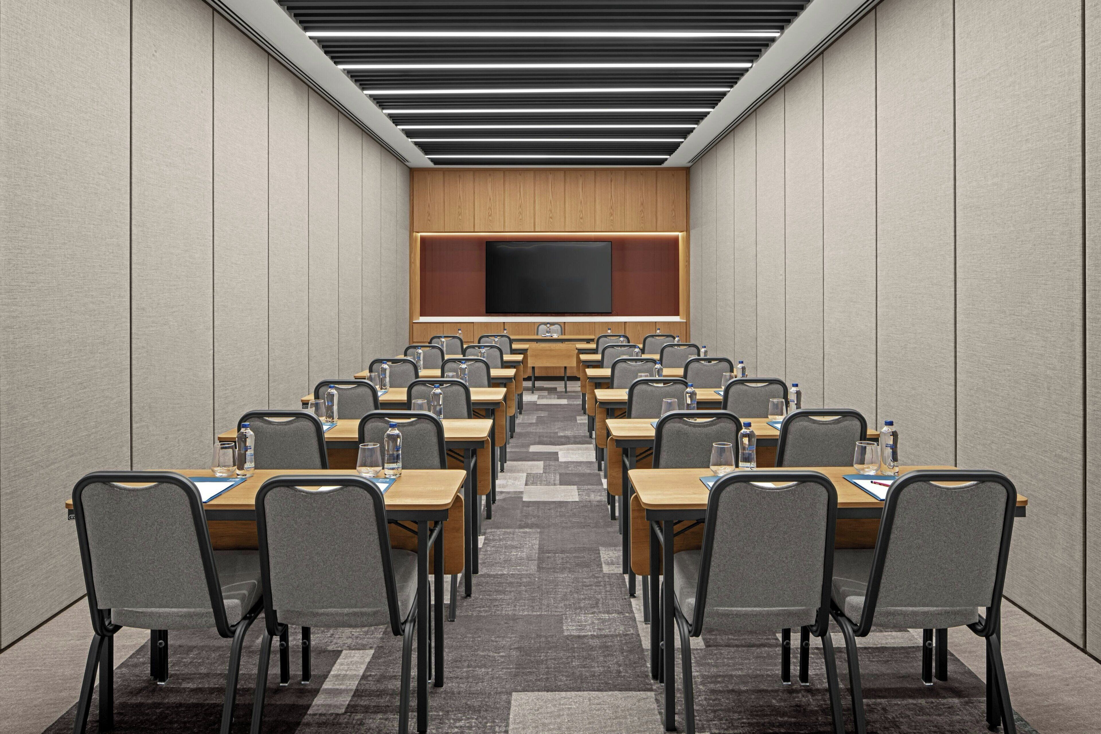 A conference room with a long table, multiple chairs, a large screen, and a wooden panel wall.