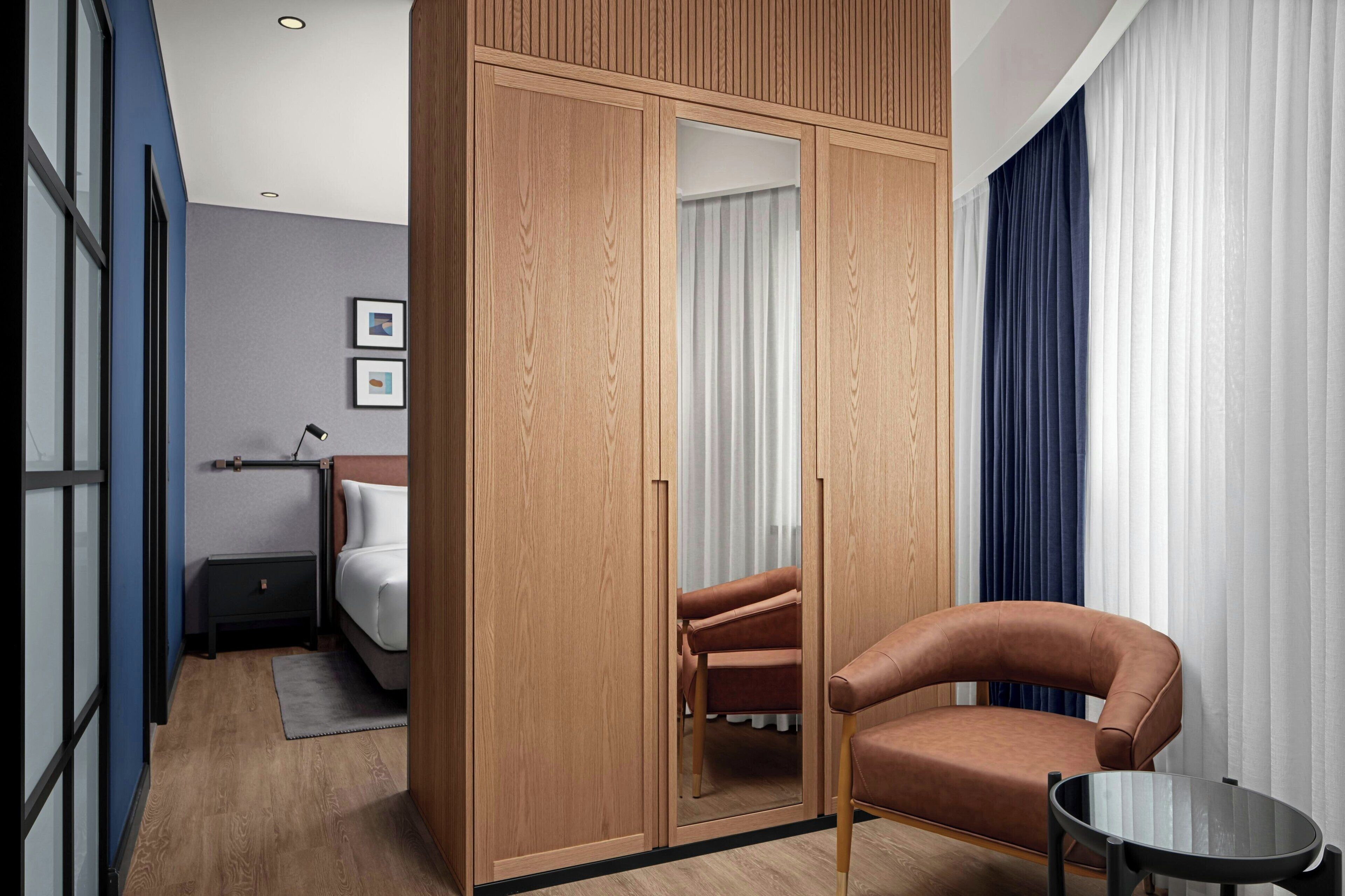 Una habitación de hotel moderna con un armario de madera, un sillón de cuero, una mesita redonda y una cama con cabecera blanca.