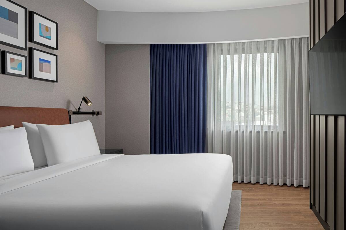 Une chambre d’hôtel moderne avec un grand lit, une table de chevet, une lampe et une fenêtre avec des rideaux légers.