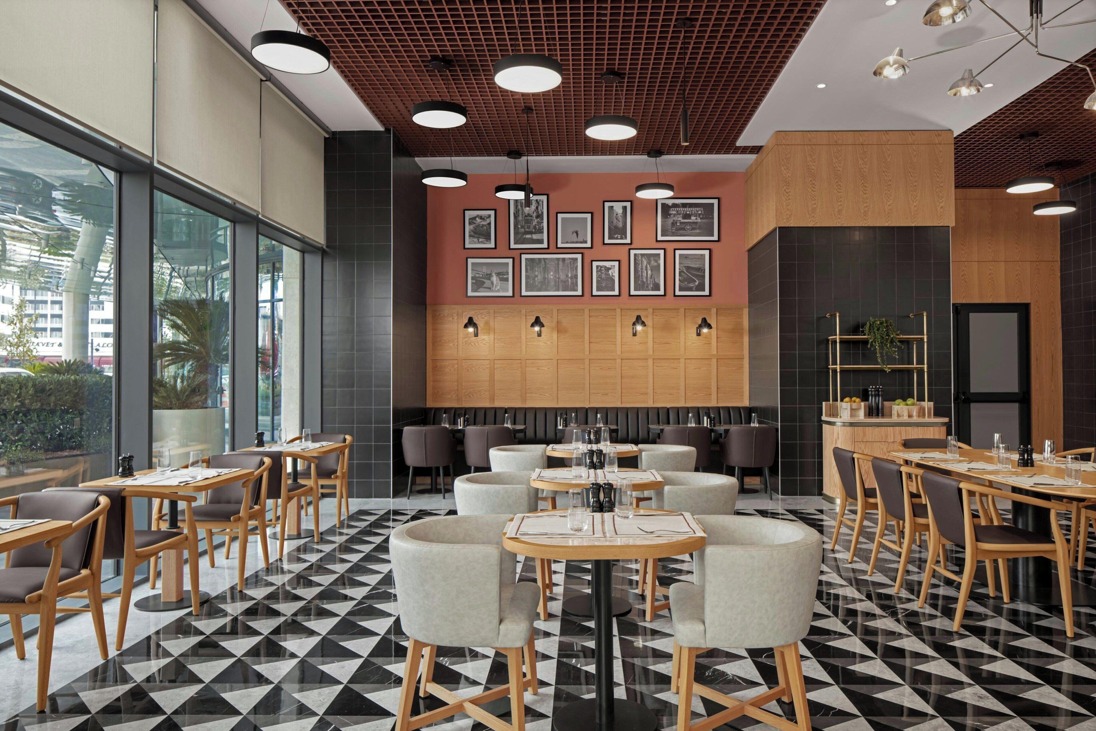 A modern restaurant with a black and white checkered floor, wooden tables and chairs, and a wall decorated with framed pictures.