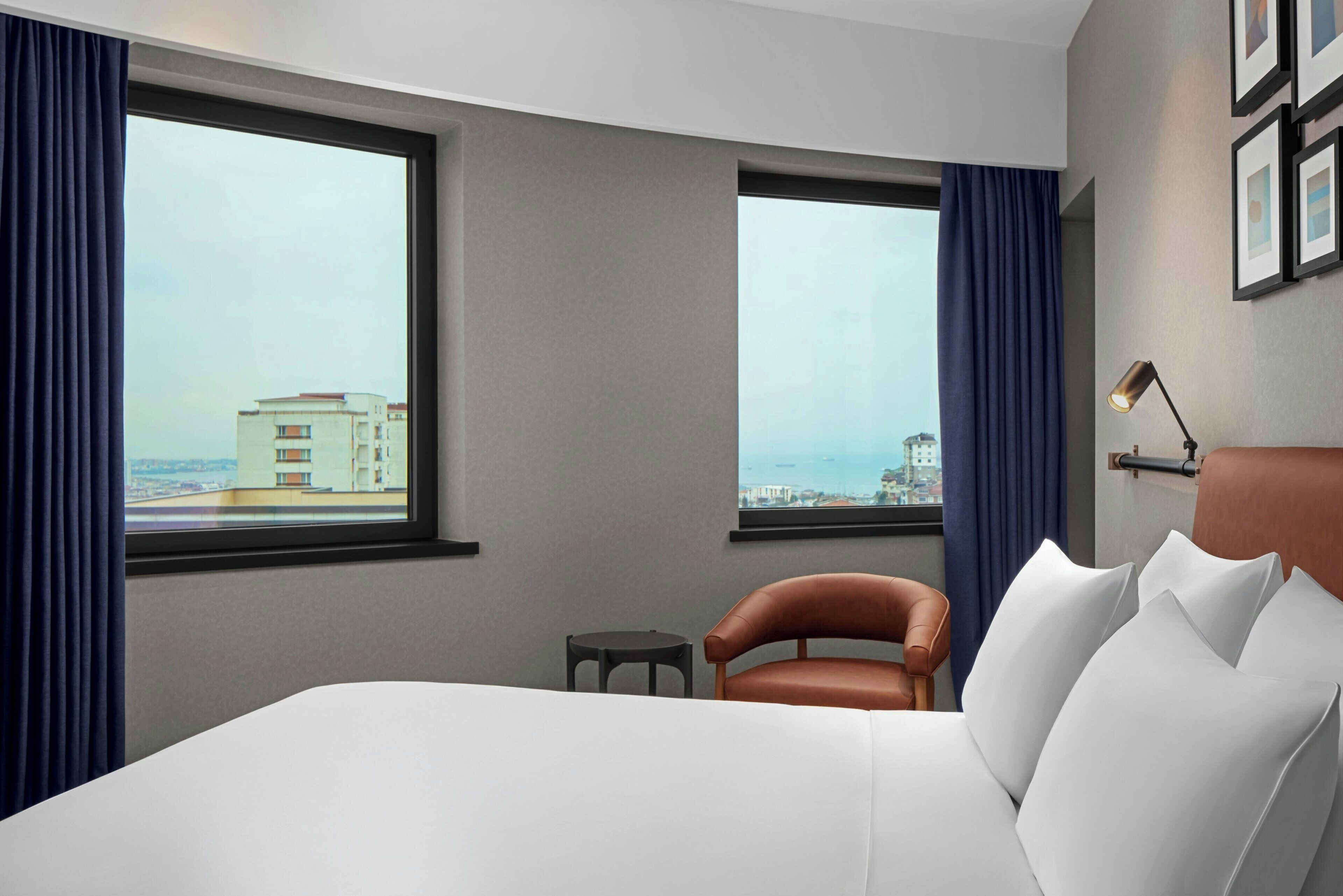 A hotel room with a large bed, two windows with curtains, a small table, a chair, and a lamp.