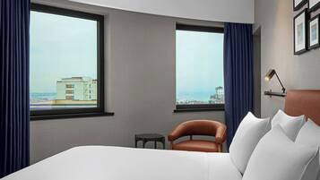 A hotel room with a large bed, two windows with curtains, a small table, a chair, and a lamp.