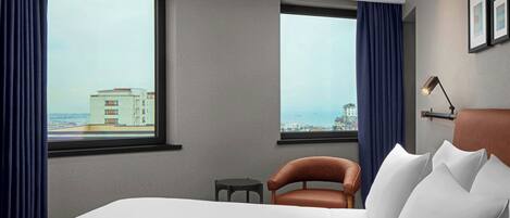 Una habitación de hotel con una cama grande, dos ventanas con cortinas, una mesita pequeña, una silla y una lámpara.