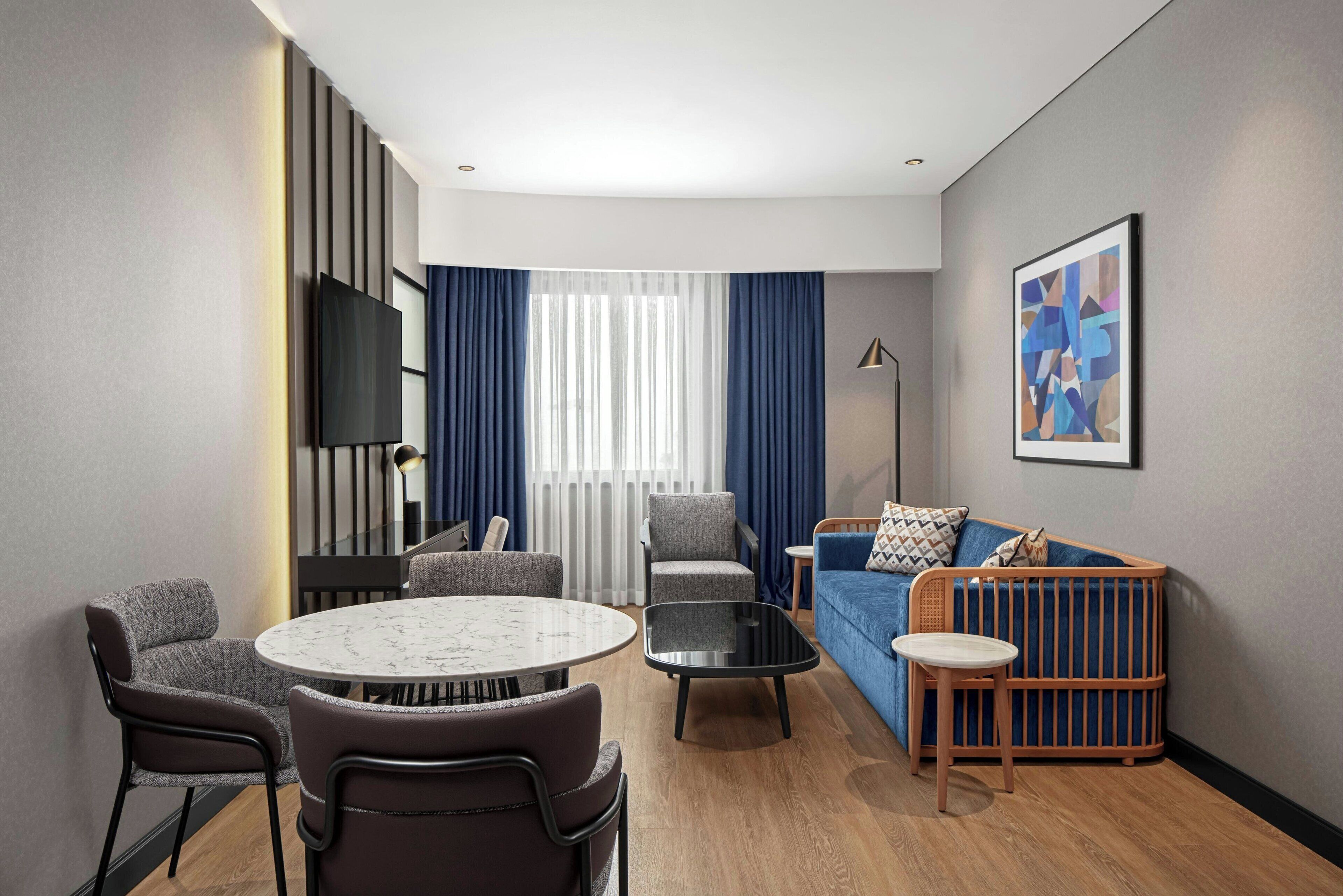 Une chambre d’hôtel moderne avec une table à manger ronde, deux fauteuils, un canapé, un berceau, une télévision et une grande fenêtre avec des rideaux.