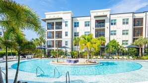 A swimming pool surrounded by lounge chairs and umbrellas, with a multi-story building in the background. - Tropical Dream 2BR/2BA w./ Pool, Gym, and Theater (Naples)