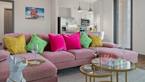 Un salon moderne avec un canapé d’angle rose, une table basse en verre et une cuisine en arrière-plan.