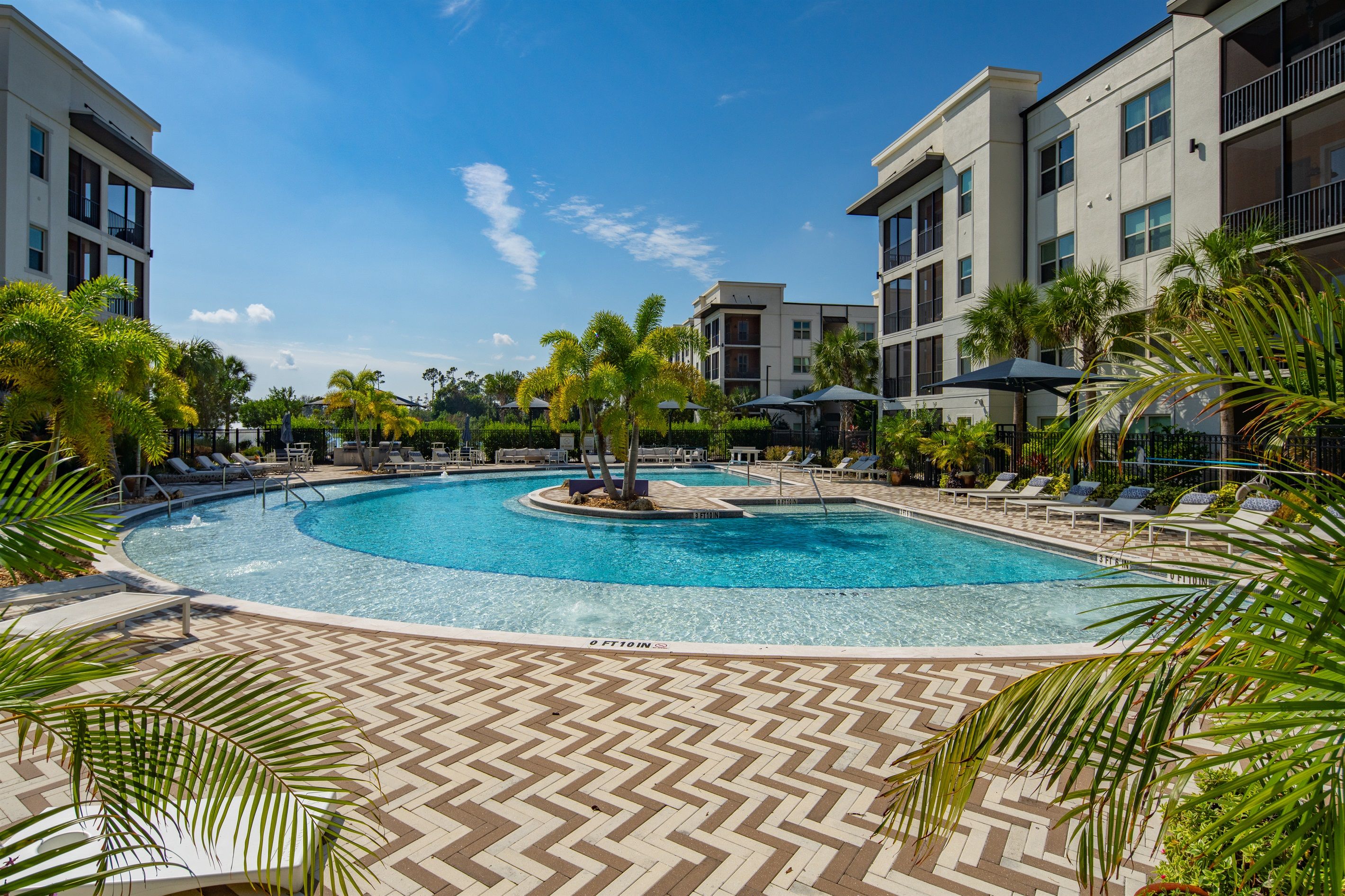 A swimming pool with lounge chairs and palm trees, surrounded by multi-story buildings.