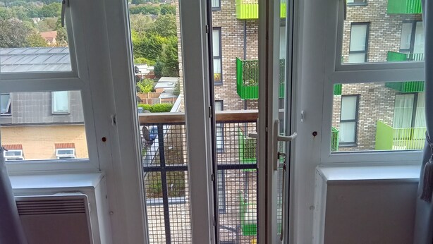A room with a large sliding glass door leading to a balcony. The balcony has a green railing and a view of a brick building with multiple windows. - Cosy London Apartment Retreat - Colindale (London)