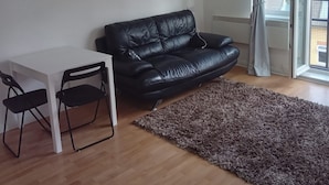 A living room with a black leather sofa, a white table, two black chairs, a brown shag rug, and a sliding glass door with a view of greenery outside. - Cosy London Apartment Retreat - Colindale (London)