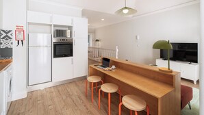 Une cuisine moderne avec des meubles blancs, un îlot central en bois doté de tabourets, un ordinateur portable et un téléviseur.