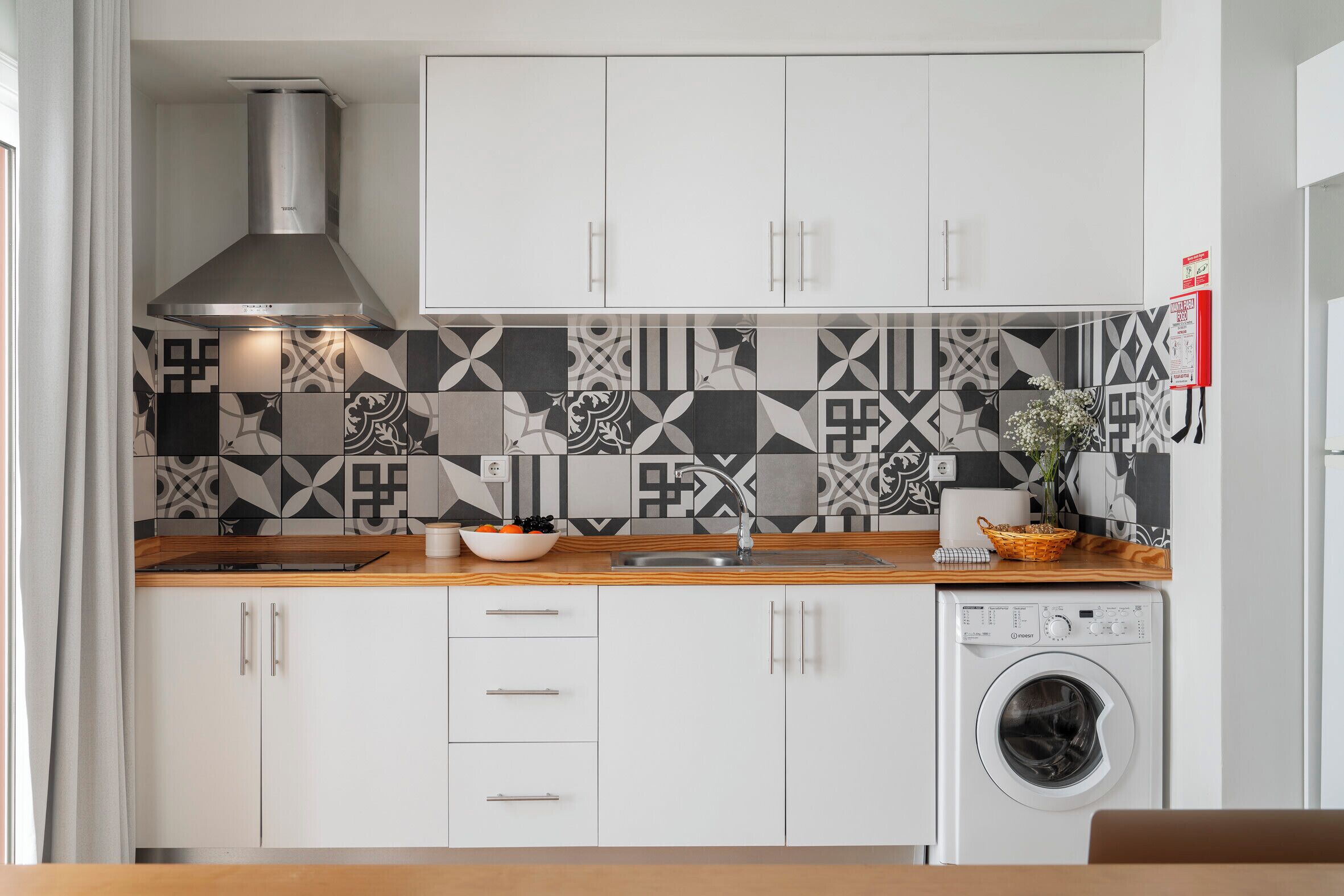 A modern kitchen with white cabinets, a stainless steel range hood, a patterned backsplash, a sink, a washing machine, and a wooden countertop.