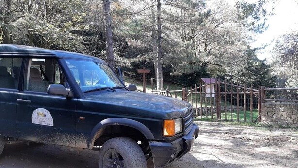 Un SUV negro estacionado en un camino de tierra, con una cerca de madera y un muro de piedra al fondo.