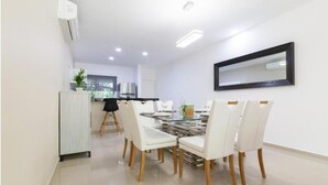 Un coin repas avec une table en verre et des chaises blanches, une cuisine à l’arrière-plan et un grand miroir sur le mur.